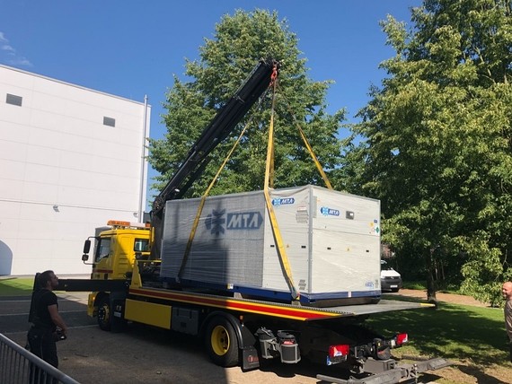 Ein LKW mit einem Kran hebt einen großen Container auf, während Menschen im Hintergrund beobachten.