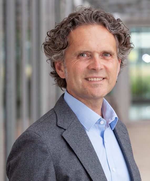 Ein Mann mit lockigem Haar, lächelnd, trägt ein Hemd und einen Anzug.