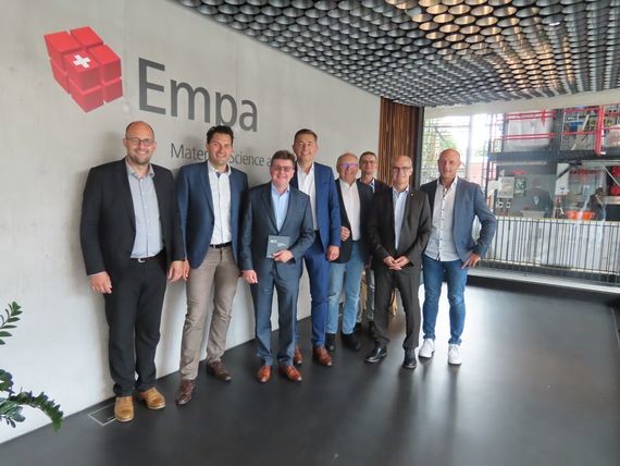 Eine Gruppe von sieben Männern steht vor einem Empa-Logo und einem modernen Bürointerieur.