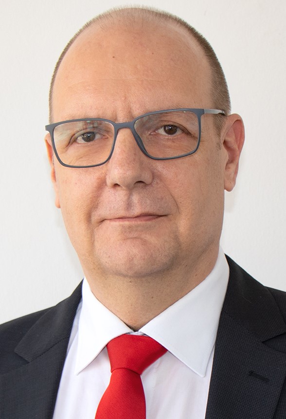 Ein Mann mit Brille, weißem Hemd und roter Krawatte steht vor einem blauen Hintergrund.