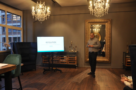 Ein Mann steht vor einer Präsentation auf einem Bildschirm in einem eleganten Raum mit Spiegel, Kronleuchtern und Holzmöbeln.