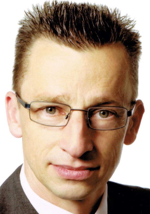 Ein Mann mit kurzen, dunklen Haaren, Brille und einem lächelnden Gesicht, der ein Hemd und eine Krawatte trägt.