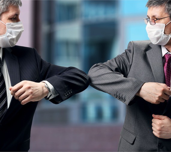 Zwei Männer in Anzügen und Mundschutz führen einen Ellbogenstoss durch.