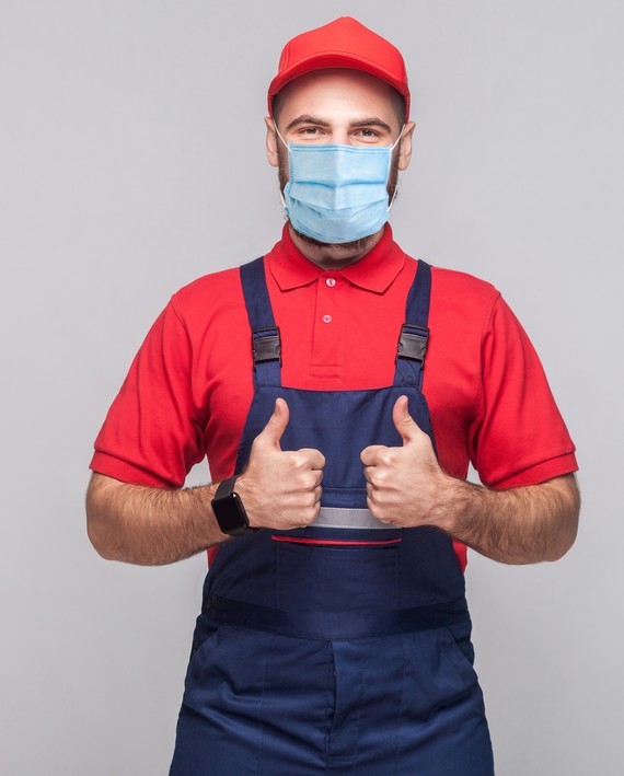 Ein Mann in rotem Hemd, blauen Overalls und einer roten Kappe zeigt Daumen hoch, während er eine Mund-Nasen-Maske trägt.