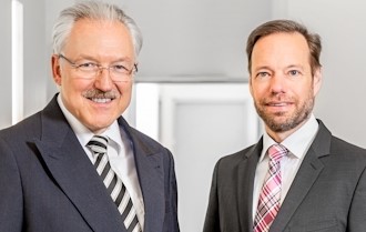 Zwei Männer in Anzügen stehen nebeneinander und lächeln.