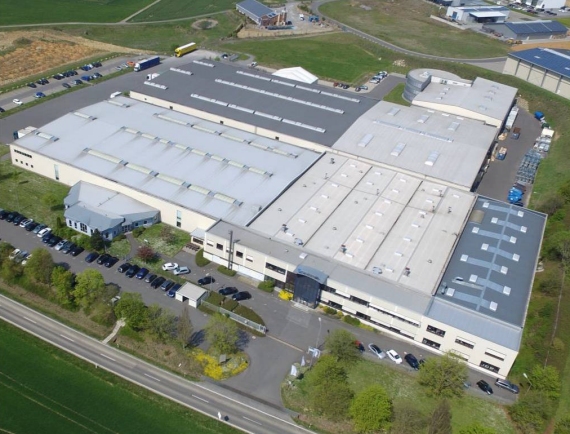 Ein Luftbild eines großen Industriegebäudes mit mehreren Gebäudeteilen, Parkplätzen und umgebendem Grünland.
