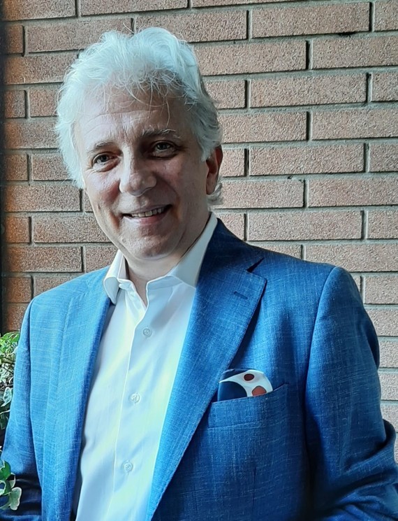 Ein Mann mit grauem Haar trägt einen blauen Anzug und steht vor einer Backsteinwand.