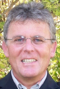 Ein Mann mit Brille und grauem Haar lächelt vor einem grünen Hintergrund.