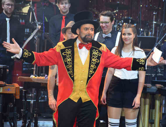 Ein Zirkusdirektor in einem auffälligen roten Umhang und Zylinder steht neben einer Zirkusdarstellerin in Uniform, während im Hintergrund weitere Personen und Instrumente zu sehen sind.