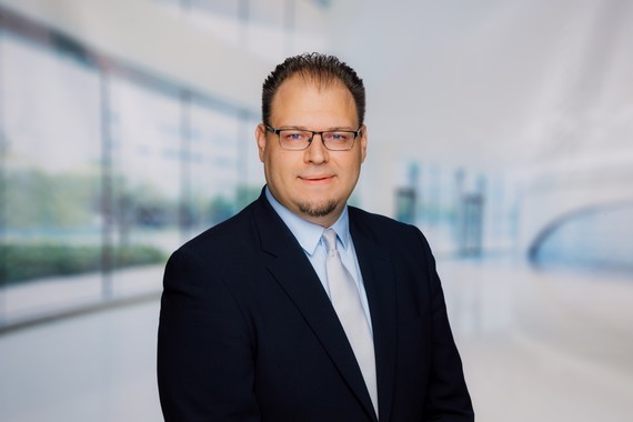 Ein Mann im Anzug und Krawatte steht vor einem unscharfen, modernen Bürohintergrund.