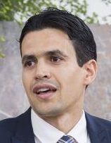 Ein Mann mit dunklem Haar und Bart trägt ein weißes Hemd und eine dunkle Krawatte, steht vor einem unscharfen Hintergrund mit grünen Pflanzen.