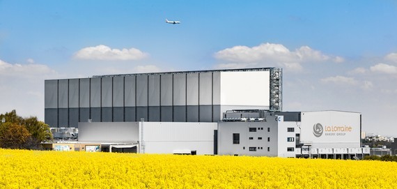 Ein großes modernes Gebäude mit einer gelben Blumenwiese im Vordergrund und einem Flugzeug am Himmel.