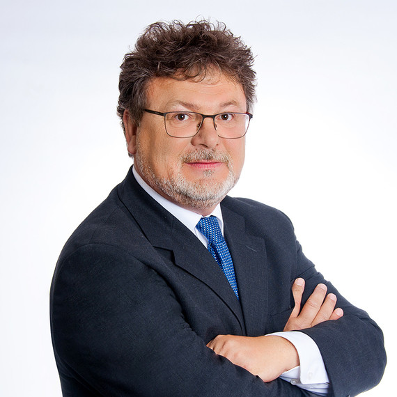 Ein Mann mit Brille, Bart und dunklem Anzug steht mit verschränkten Armen vor einem weißen Hintergrund.