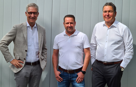 Drei Männer stehen nebeneinander lächelnd vor einer grauen Wand.