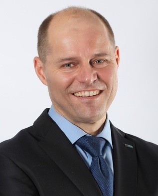 Ein Mann mit kurzen Haaren, lächelnd, trägt einen dunklen Anzug mit Krawatte.