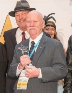 Ein Mann in Anzug und Krawatte hält einen Preis in der Hand, während im Hintergrund weitere Personen zu sehen sind.