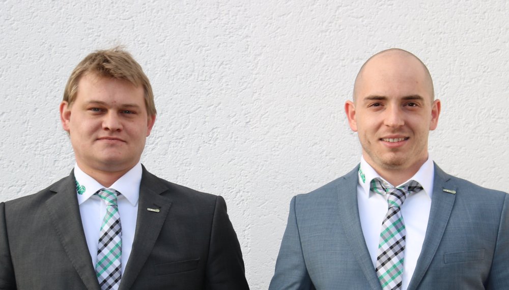 Zwei Männer in Anzügen und Krawatten stehen nebeneinander vor einem weißen Hintergrund.