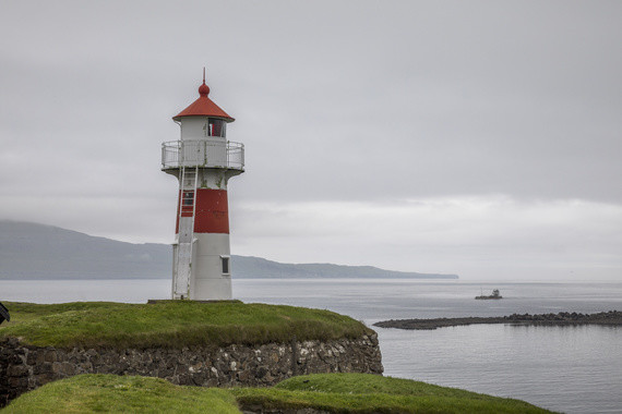Ein roter und weißer Leuchtturm steht auf einer grünen Halbinsel am Meer unter einem bewölkten Himmel.