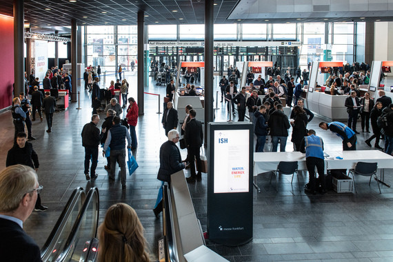 Ein belebter Veranstaltungsort mit Menschen, die sich in einem großen Raum mit moderner Einrichtung bewegen, während ein digitales Informationsdisplay im Vordergrund steht.
