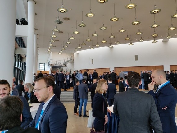 Eine Gruppe von Menschen in formeller Kleidung unterhält sich in einem großen, hellen Veranstaltungssaal.