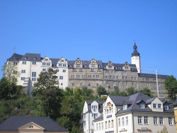 Ein großes historisches Schloss thront auf einem Hügel über einer idyllischen Stadtlandschaft mit kleineren Häusern im Vordergrund.
