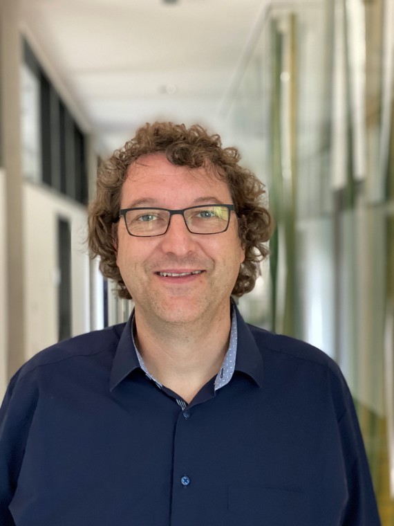 Ein Mann mit kurzen braunen Haaren und Brille trägt ein dunkelblaues Hemd und steht in einem hellen Flur.