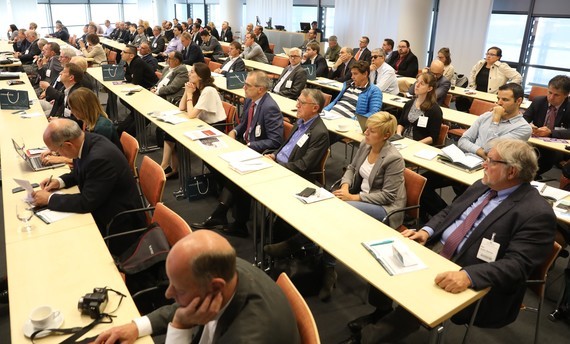 Eine Gruppe von Menschen sitzt in einer Konferenzhalle an langen Tischen und hört aufmerksam zu.