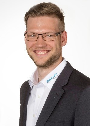 Ein Mann mit Brille, Bart und kurzen Haaren trägt ein weißes Hemd und einen dunklen Anzug mit einem Logo auf der Brust.