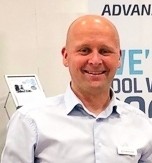 Ein Mann in einem hellblauen Hemd steht vor einem Hintergrund mit dem Text 