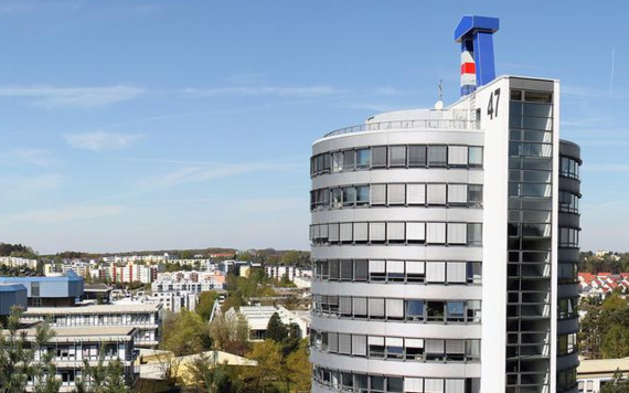 Ein modernes, rundes Hochhaus mit einem markanten Logo auf der Spitze steht im Vordergrund, während im Hintergrund eine städtische Landschaft mit weiteren Gebäuden und Bäumen zu sehen ist.