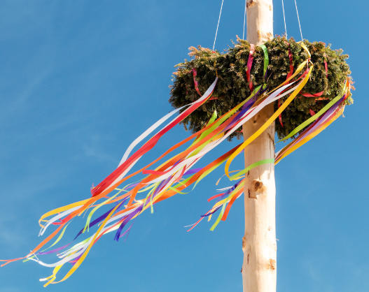 Ein traditioneller Maibaum mit einem Kranz aus Eichenlaub und bunten Bändern, der gegen einen blauen Himmel steht.