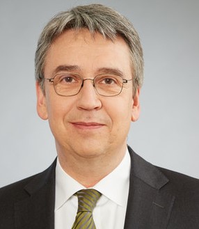 Ein Mann im Anzug mit Krawatte und Brille schaut direkt in die Kamera.