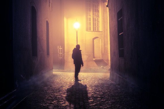 Eine einsame Person steht in einer nebligen, beleuchteten Gasse bei Nacht.