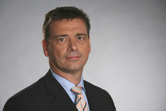 Ein Mann mit kurzem dunklem Haar trägt einen dunklen Anzug, ein weißes Hemd und eine gemusterte Krawatte, vor einem neutralen Hintergrund.