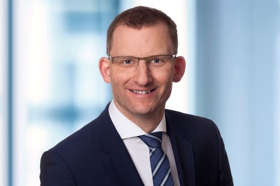 Ein Mann in Anzug und Krawatte steht vor einem unscharfen Hintergrund.