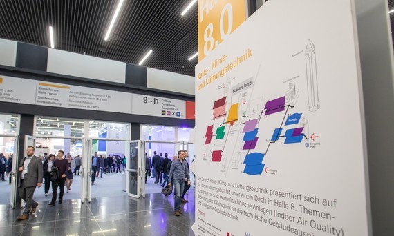 Eine Präsentationswand mit Diagrammen und Text steht im Hintergrund eines belebten Messestandes, wo Menschen diskutieren und sich bewegen.