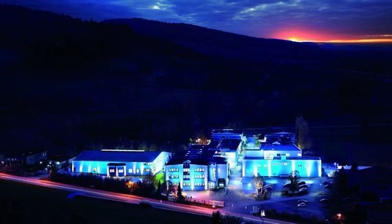 Ein beleuchtetes Gebäude mit blauen Lichtern steht im Vordergrund, während im Hintergrund ein abendlicher Himmel mit einem orangefarbenen Sonnenuntergang über einer dunklen Landschaft zu sehen ist.