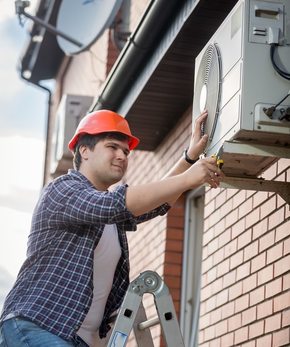 Ein Mann in einem orangefarbenen Helm und Schutzausrüstung montiert ein Gerät an einer Hauswand, während er auf einer Klappleiter steht.