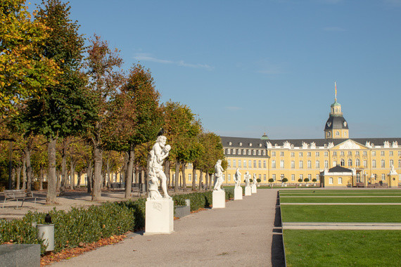Ein gepflegter Park mit einer Allee von Bäumen, Skulpturen und einem großen, klassizistischen Gebäude im Hintergrund.
