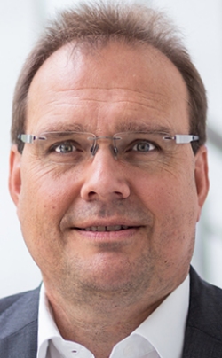 Ein Mann mit Brille und kurzem Haar lächelt in die Kamera, im Hintergrund ist ein unscharfer grüner Hintergrund zu sehen.