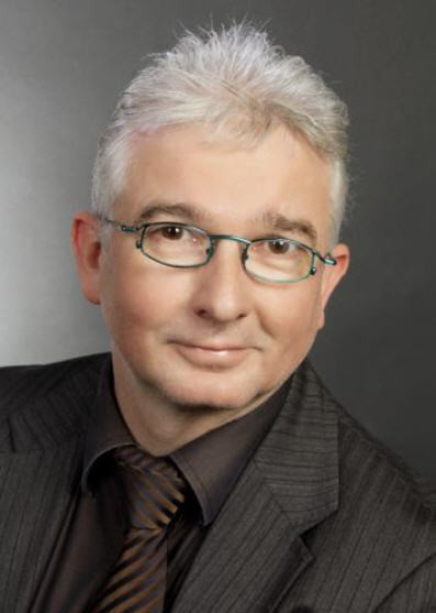 Ein Mann mit grauen Haaren, Brille, Hemd und Krawatte steht vor einem dunklen Hintergrund.