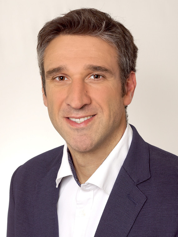 Ein Mann mit dunklem Haar und Bart trägt ein weißes Hemd und einen dunklen Blazer.