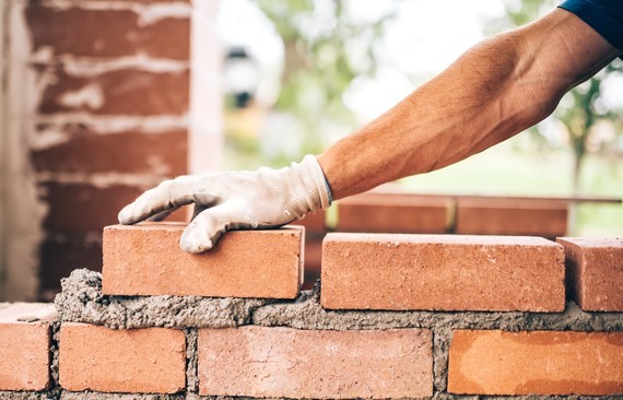 Eine Hand in einer Arbeitshandschuh legt einen Ziegel auf einen Backsteinmauerbau.