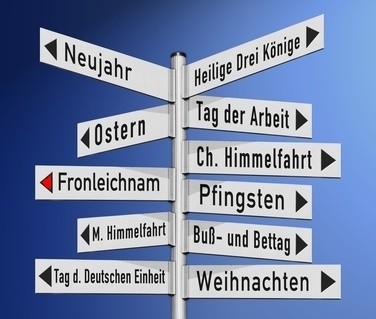 Ein Schild mit mehreren Pfeilen zeigt Richtungen zu verschiedenen Feiertagen wie Neujahr, Heilige Drei Könige, Ostern, Tag der Arbeit, Fronleichnam, Pfingsten, M. Himmelfahrt, Buß- und Bettag, Tag der Deutschen Einheit und Weihnachten.