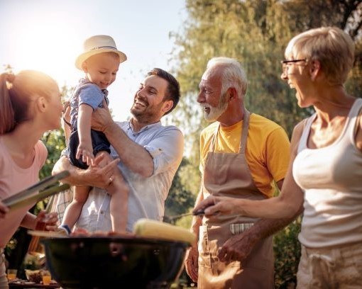 Eine glückliche Familie feiert gemeinsam im Freien, wobei ein Mann ein Kind hochhält und andere Mitglieder im Hintergrund grillen und lachen.