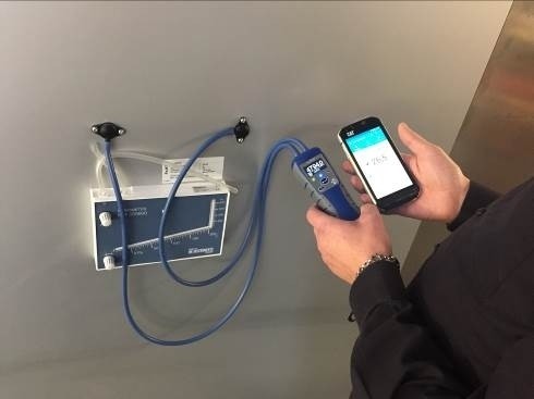 Eine Person hält ein Smartphone mit einer App in der Hand und ist dabei, einen blauen Kabelanschluss an einem Gerät an der Wand zu überprüfen.