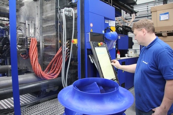 Ein Mann in blauem Hemd steht in einer Fabrikhalle und bedient ein digitales Bedienfeld neben einem großen blauen Maschinenaggregat.
