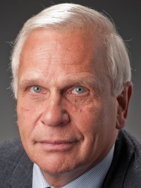 Ein älterer Mann mit grauem Haar und ernstem Gesichtsausdruck, gekleidet in ein Hemd und eine Jacke.