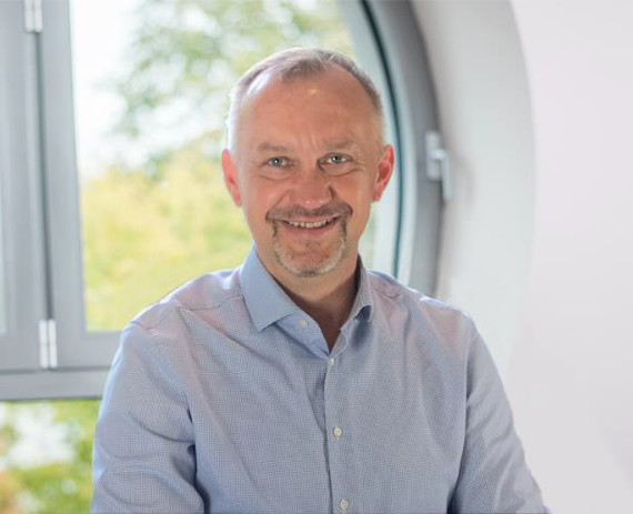 Ein Mann mit kurzen Haaren und Bart trägt ein hellblaues Hemd und sitzt vor einem Fenster mit grüner Außenansicht.