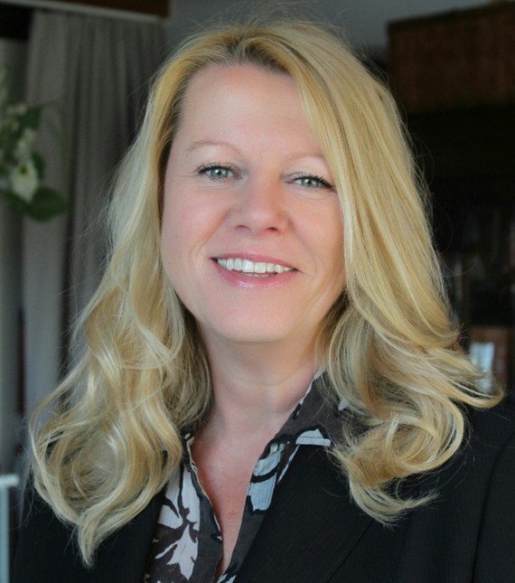 Eine Person mit blonden, lockigen Haaren und einem Lächeln im Vordergrund, im Hintergrund ist ein unscharfer Raum mit einer Pflanze zu sehen.